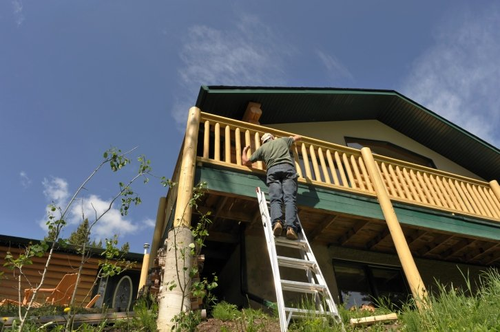 Person on ladder installing wooden railing on house deck against blue sky