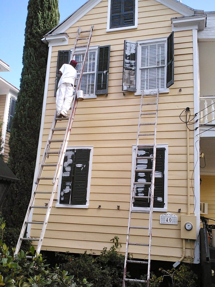 Painter in white clothes climbing ladder to paint tan house exterior with shutters