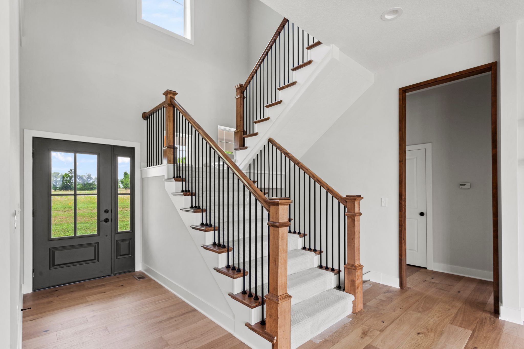 Modern foyer with wooden staircase, black metal railings, and glass entrance doors overlooking green fields